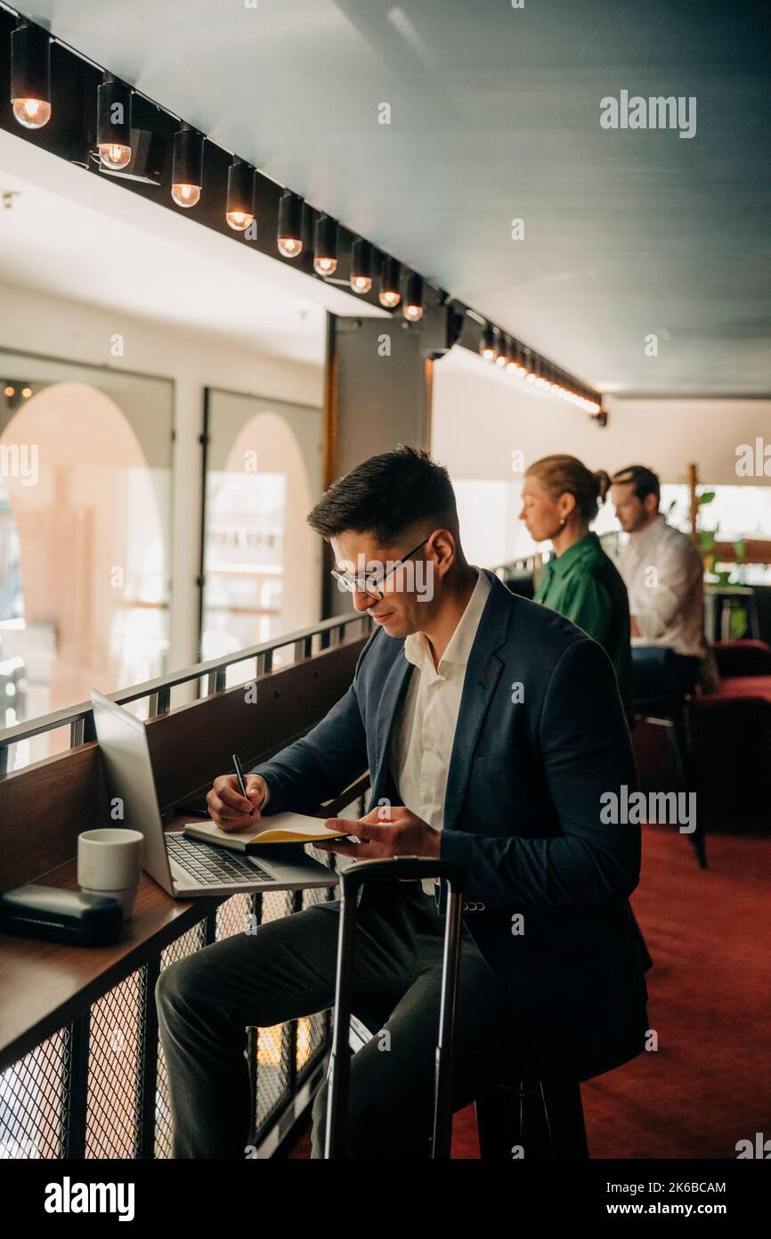 Uomo d'affari che scrive in agenda mentre si siede con il computer portatile presso l'hotel Foto Stock