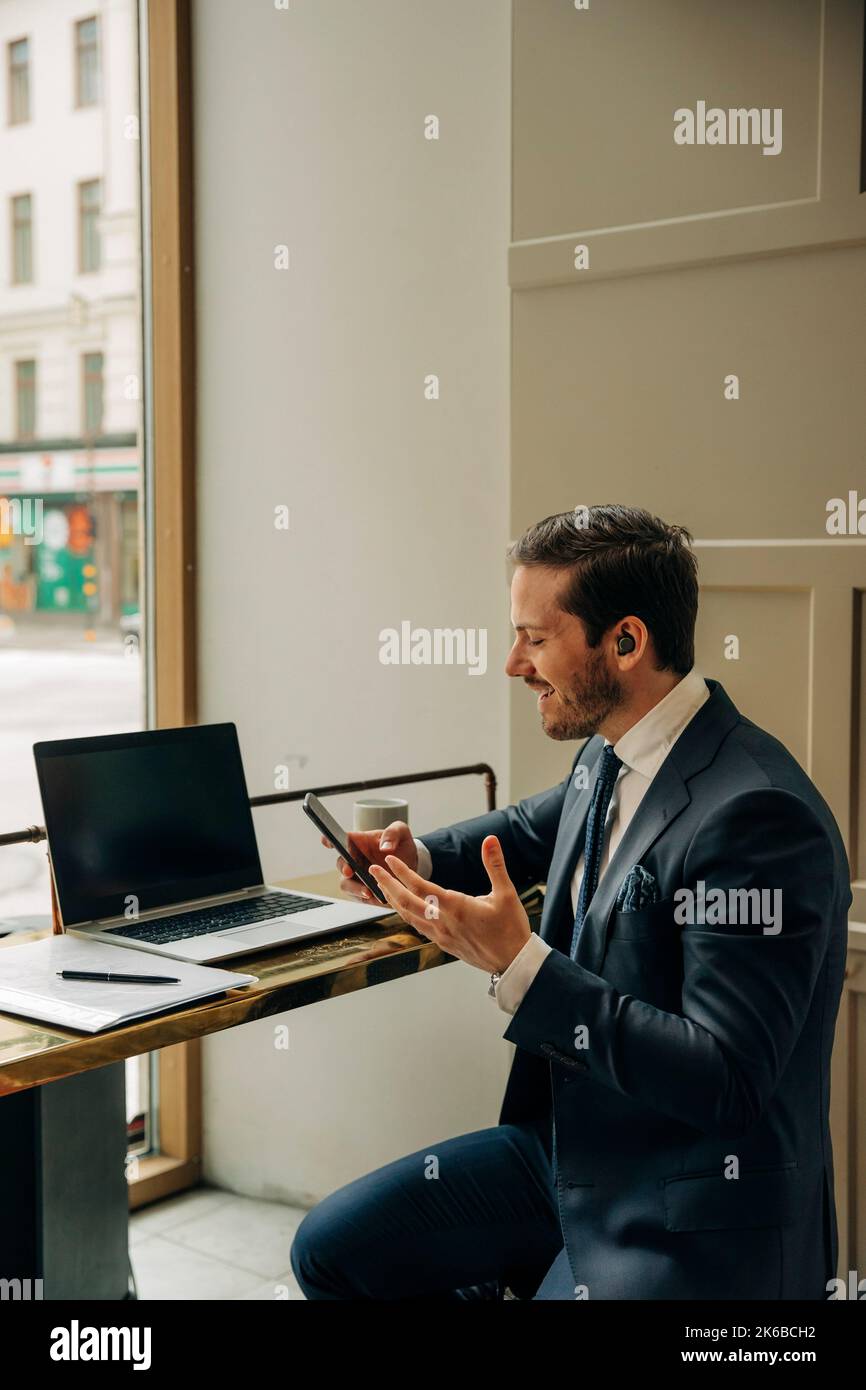 Uomo d'affari sorridente che tiene il telefono cellulare mentre parla con cuffie auricolari wireless in ufficio Foto Stock