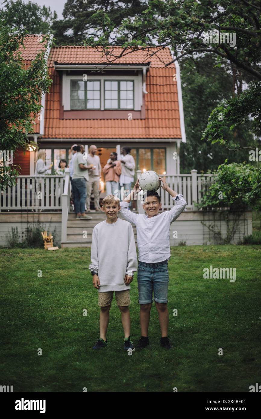 Ragazzi giocosi con palla da calcio in piedi nel cortile posteriore con la famiglia sul portico sullo sfondo Foto Stock