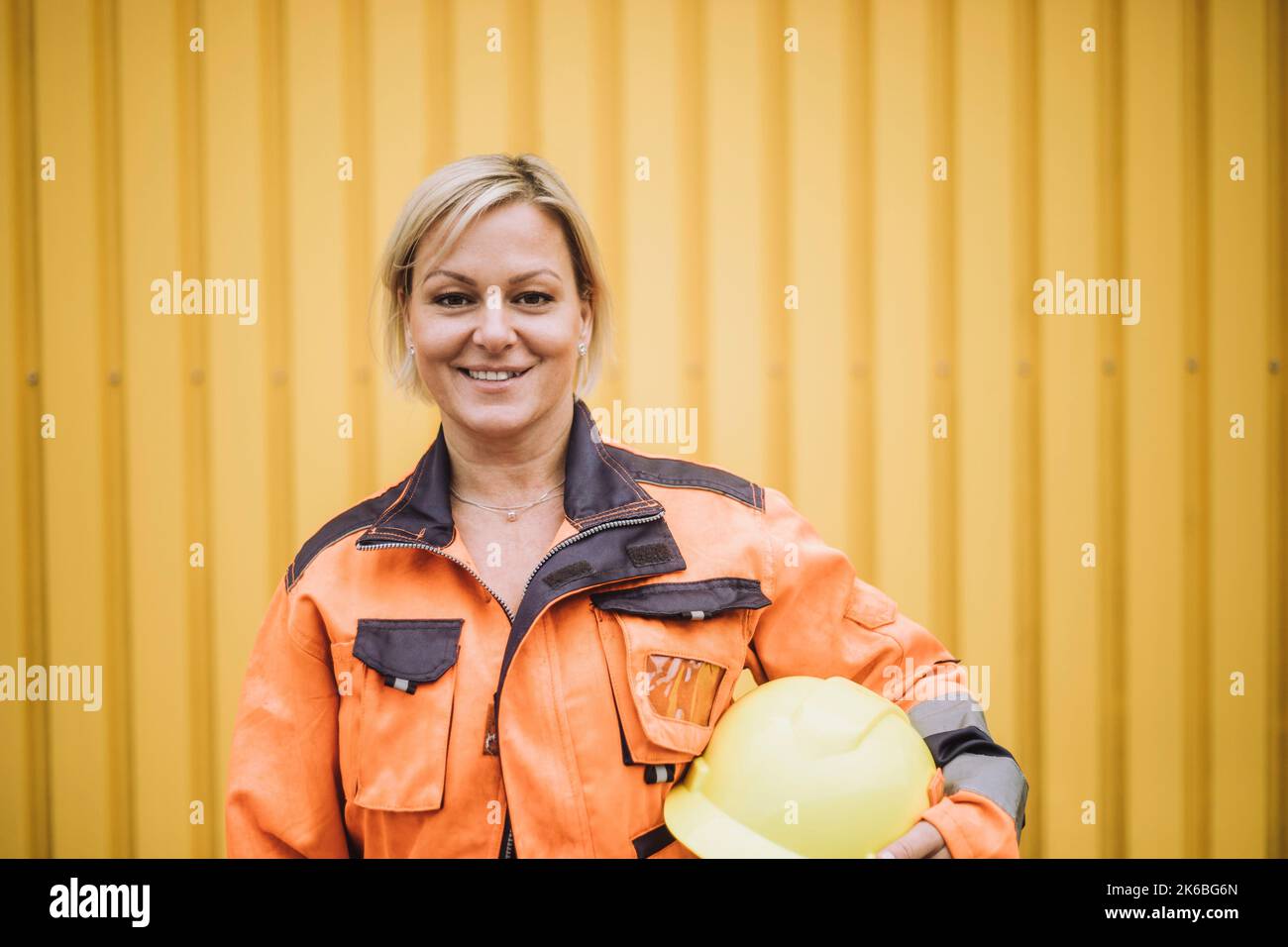 Ritratto di felice operatrice con elmetto contro muro di metallo giallo Foto Stock