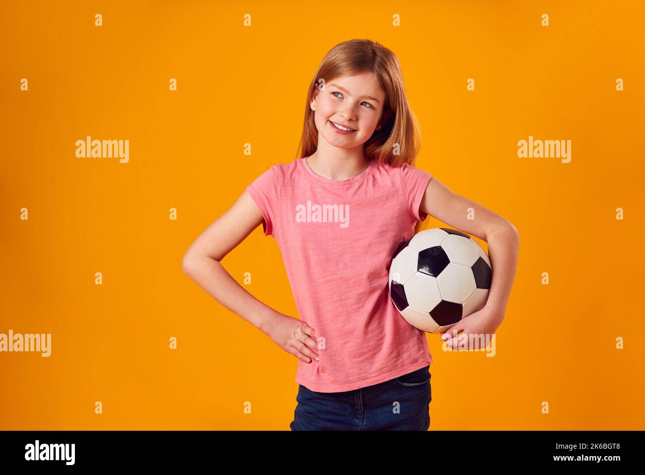 Studio Shot di Young Girl Holding palla da calcio sotto il braccio contro sfondo giallo Foto Stock