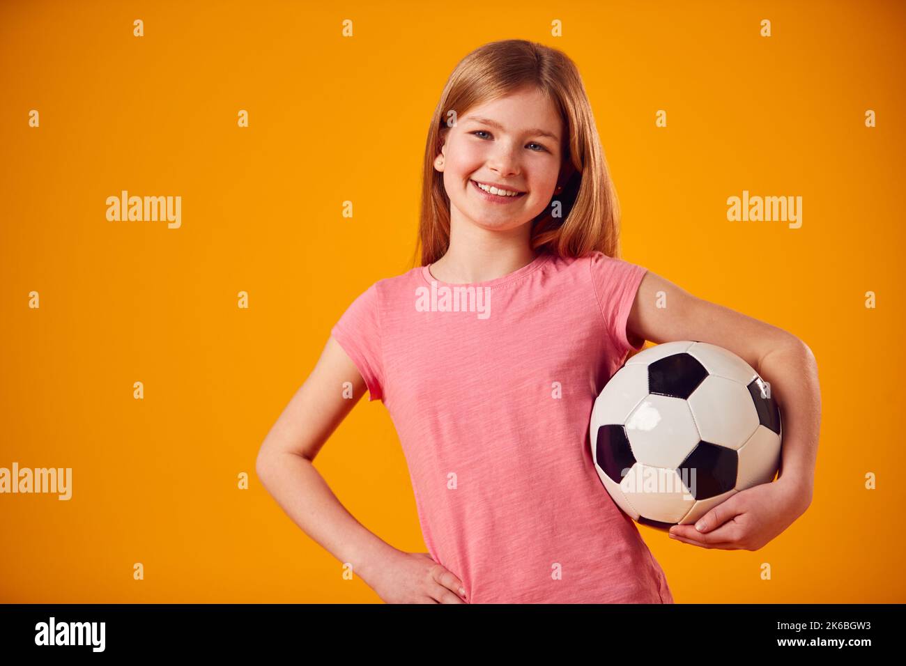 Studio Shot di Young Girl Holding palla da calcio sotto il braccio contro sfondo giallo Foto Stock