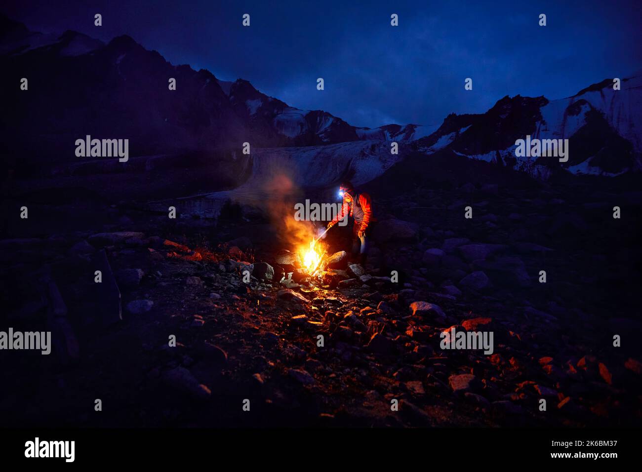Uomo turistico in giacca rossa con lampada frontale vicino al falò del campo nelle montagne innevate sotto il cielo notturno con le stelle. Foto Stock