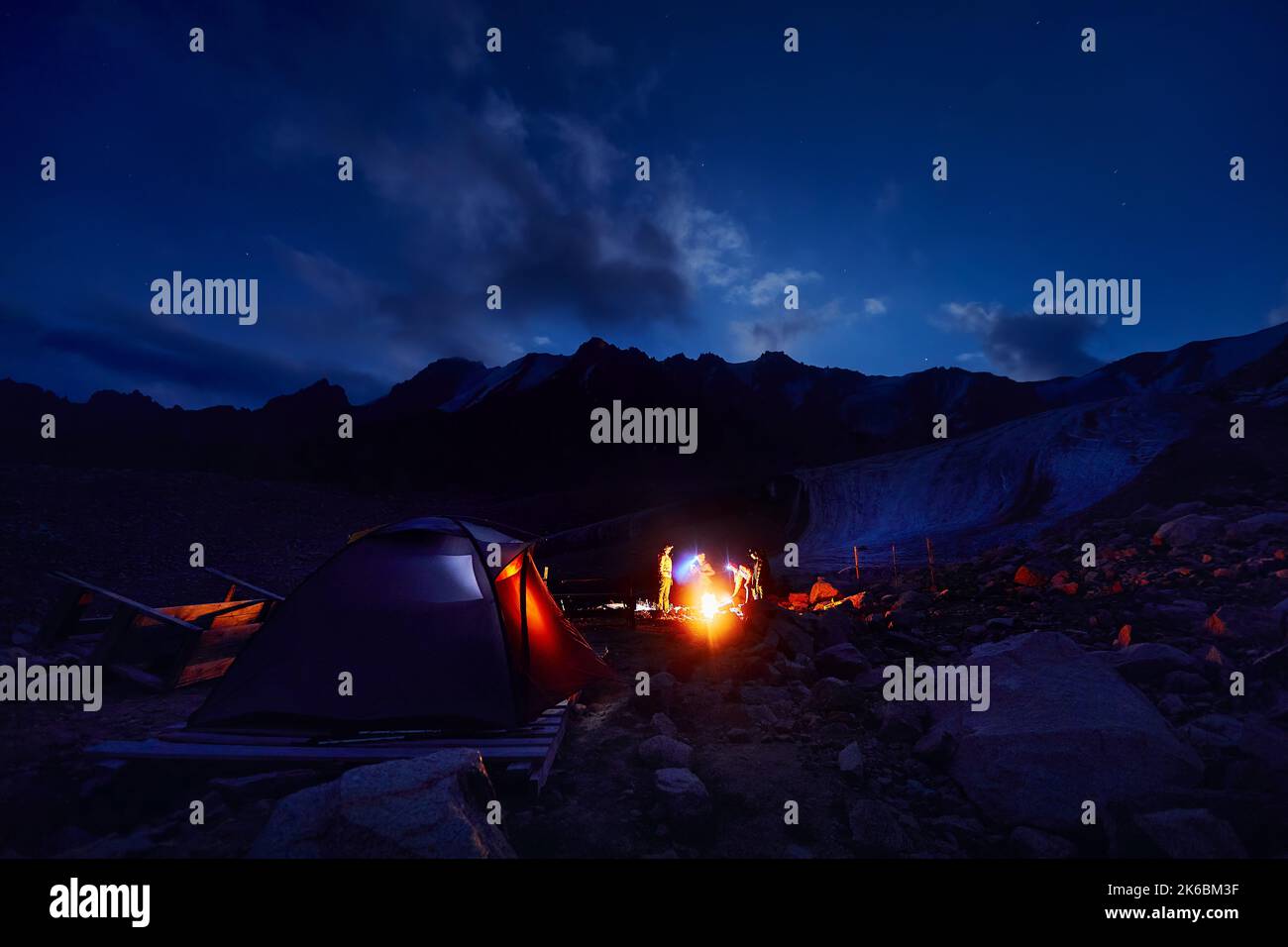 Turisti con fari vicino tenda campo illuminato vicino falò nelle montagne di neve sotto il cielo notturno con stelle. Foto Stock