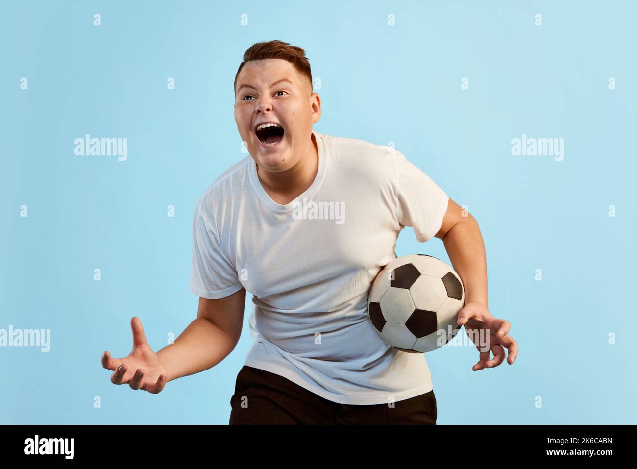 Tifoso di calcio. Ragazzo giovane eccitato, modello sovrappeso in camicia bianca con palla da calcio gridando isolato su sfondo blu studio. Sport, emozioni, gioventù Foto Stock