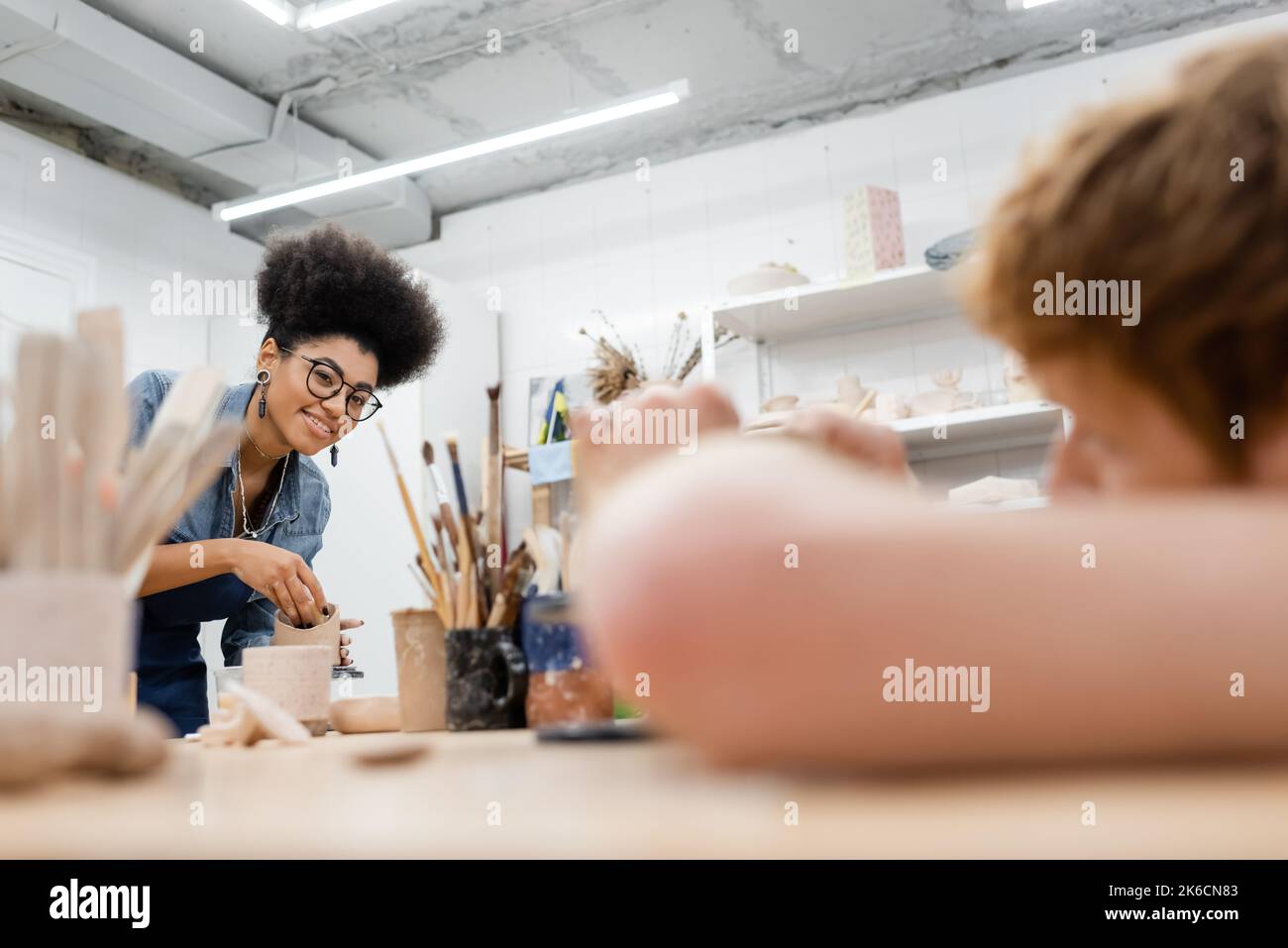 Donna afroamericana sorridente che tiene l'argilla vicino gli attrezzi sfocati ed il ragazzo in officina dell'argilla, immagine di scorta Foto Stock