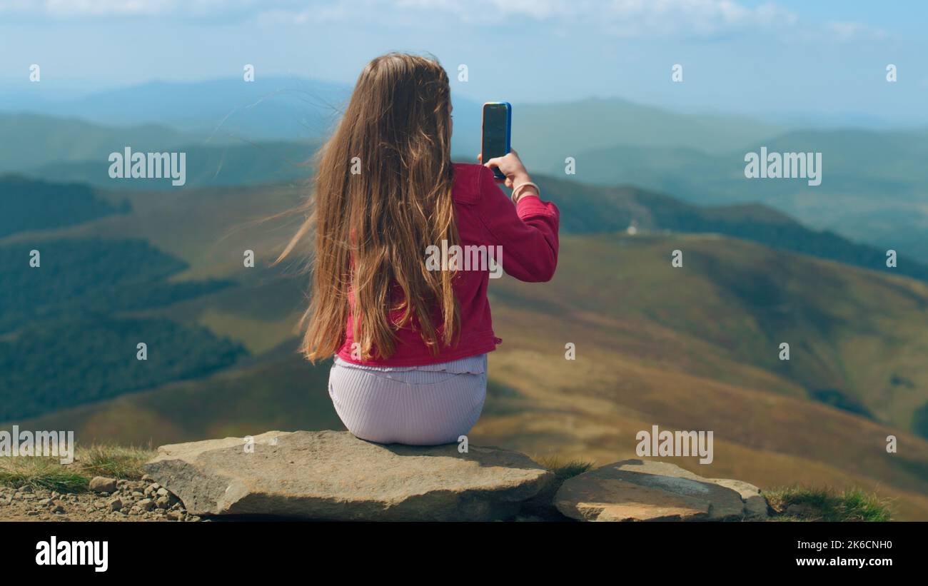 Ragazza giovane filmare il paesaggio di belle colline valle tra le montagne al telefono, seduto sulla pietra e facendo contenuti creativi in natura, trascorrere il tempo libero all'aperto. Vista posteriore. Foto Stock