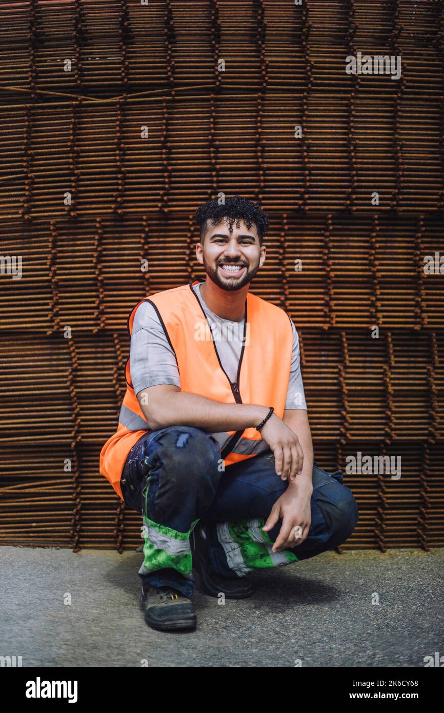 Ritratto completo di giovane lavoratore di costruzione felice accovacciato in loco Foto Stock