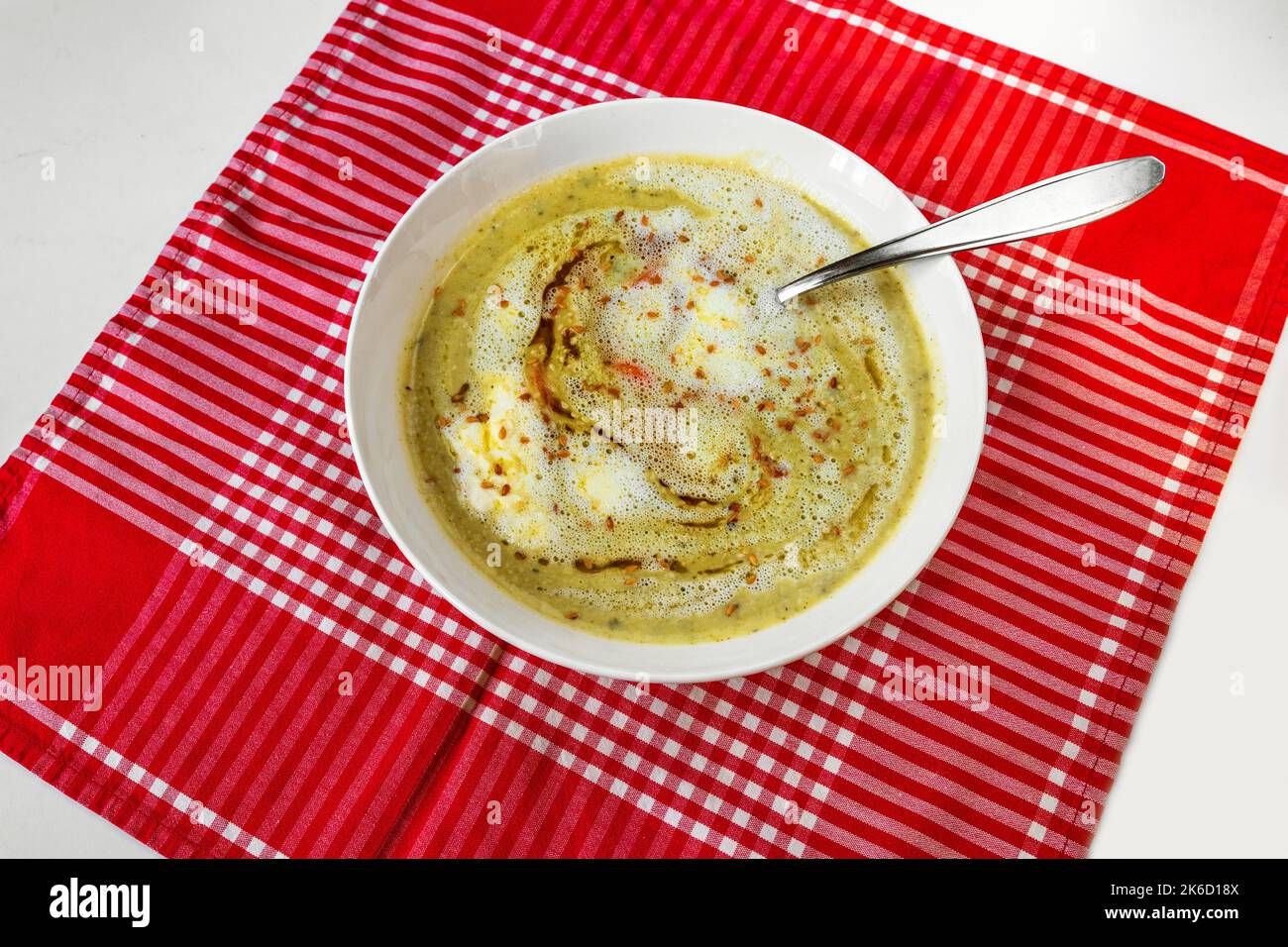 Zuppa di broccoli con semi di sesamo, olio di cumino nero e crema in piatto bianco su tavolo a scacchi rosso su fondo bianco. Foto Stock