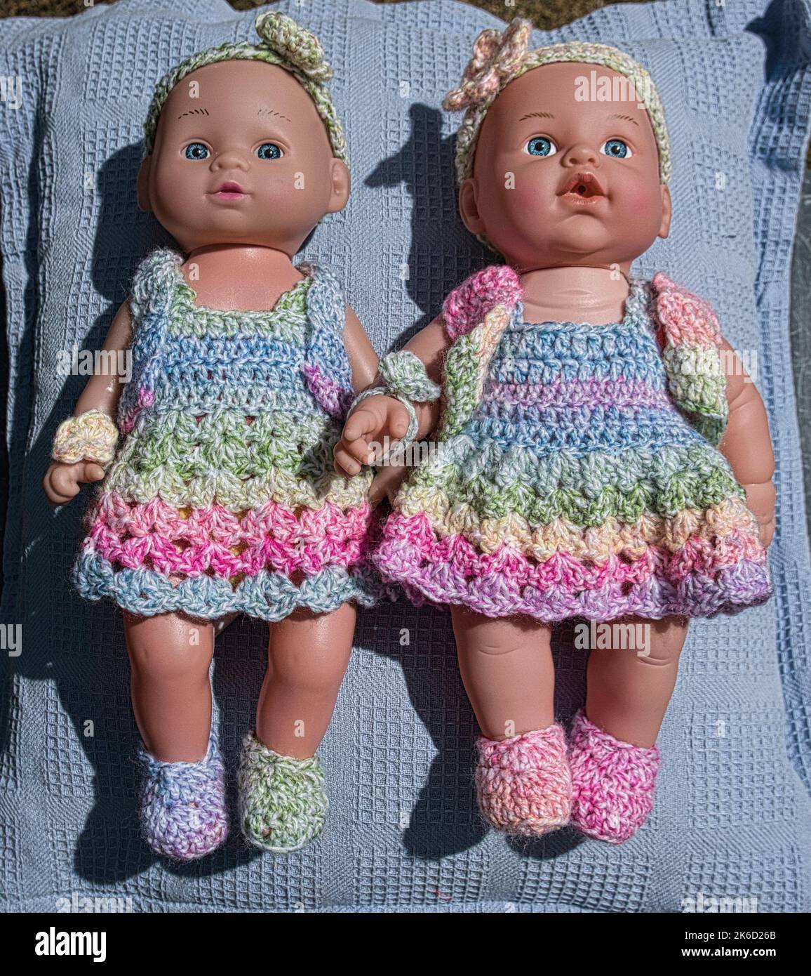 Primo piano di due bambole di bambina identiche in cappelli, abiti e bootee lavorati a maglia identici che si trovano l'una accanto all'altra e che si immergono nel sole Foto Stock