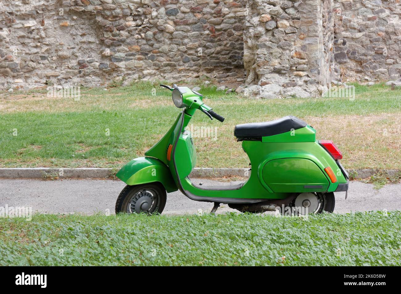 Scooter verde parcheggiato su una passerella tra due aree di erba accanto a un antico muro di pietra esterno Foto Stock