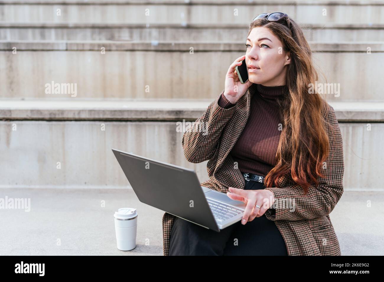 Donna in outerwear con netbook guardare lontano e avere conversazione con lo smartphone mentre si siede a pochi passi vicino a zero rifiuti Cup e lavorare in città strada Foto Stock