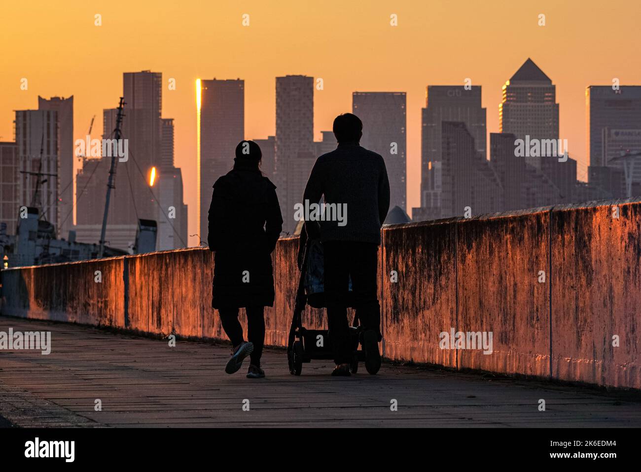 Giovane coppia con bambino che cammina lungo il Tamigi al tramonto con i grattacieli di Canary Wharf sullo sfondo, Londra, Inghilterra, Regno Unito, Regno Unito Foto Stock