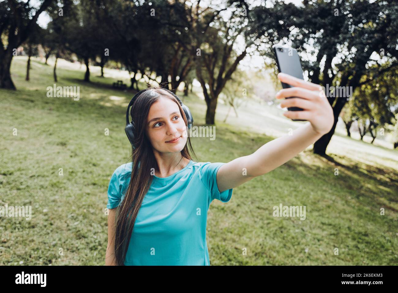 Ragazza giovane sorridente che indossa una t-shirt e cuffie turchesi, che porta un selfie con il cellulare in uno spazio naturale Foto Stock