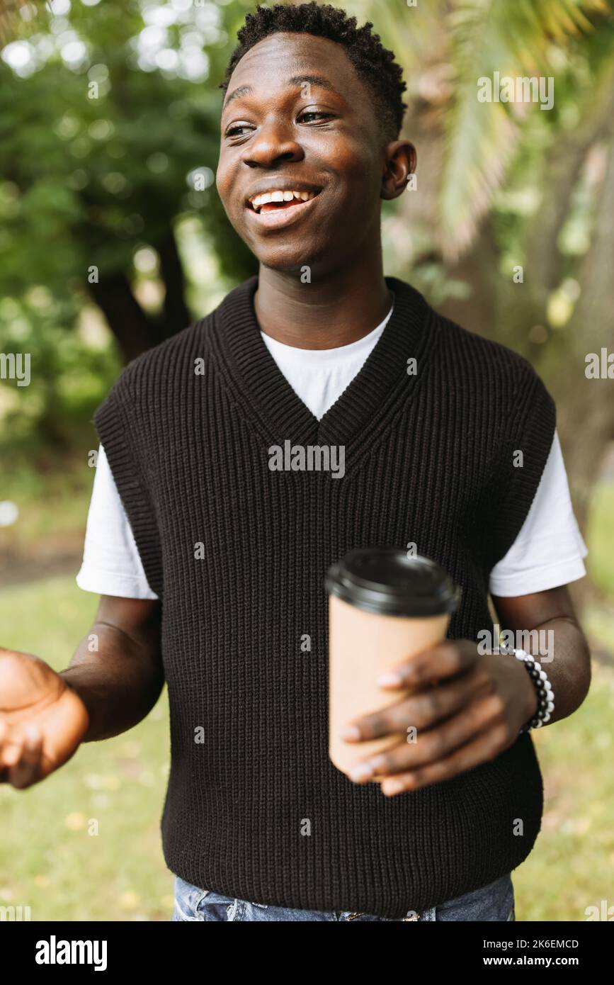 Giovane afroamericano sorridente con per andare caffè tazza sulla strada all'aperto. Giovane studente nero in pausa caffè nel parco Foto Stock