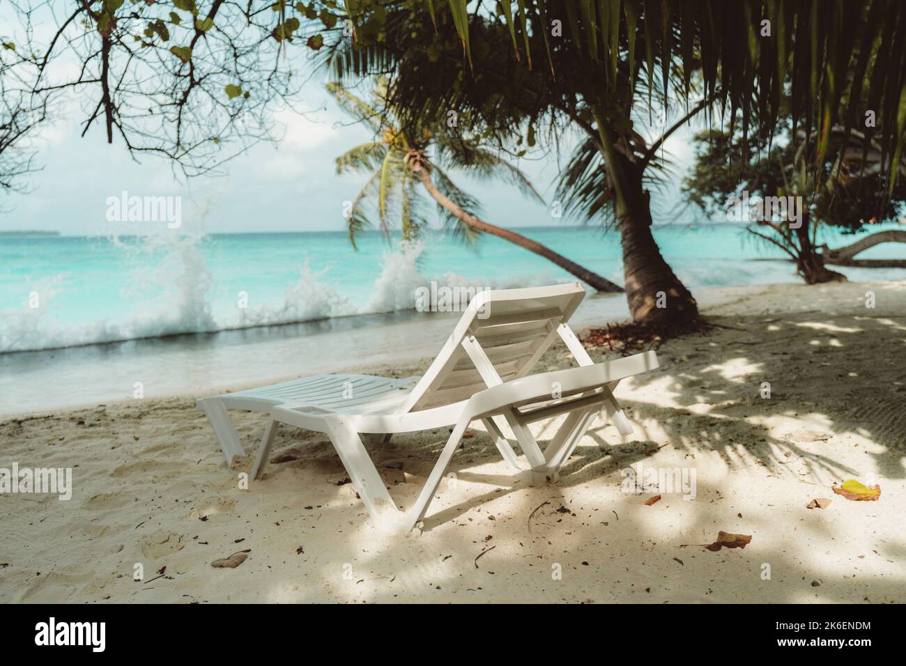 Vista di un divano letto di fronte all'oceano in un lussuoso resort delle Maldive in una calda giornata di sole circondato da palme; una sedia reclinabile in plastica sulla sabbia di corallo Foto Stock