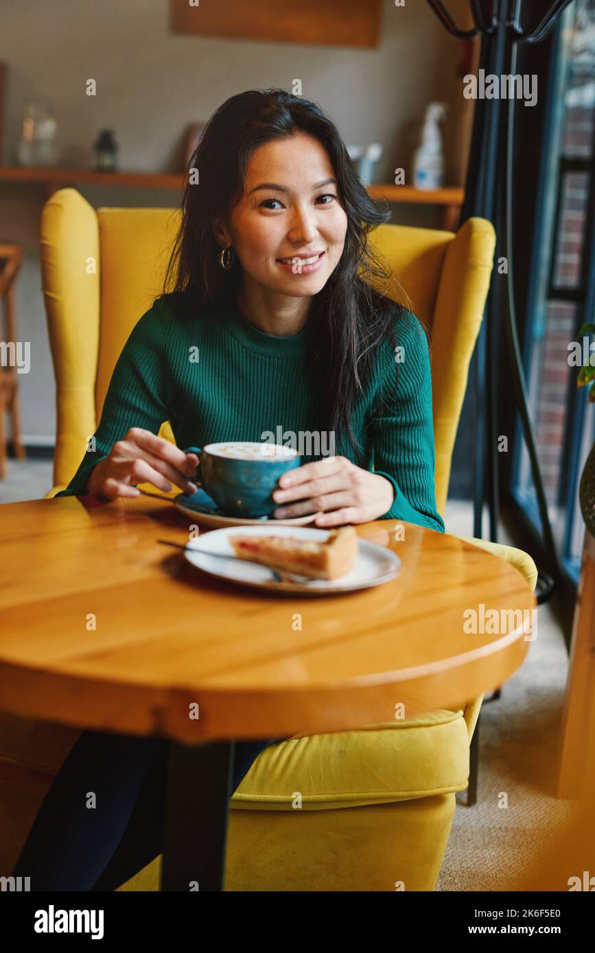 Ritratto di giovane asiatico affascinante, bella donna brunette bere caffè con torta di mele a tavola, seduta in accogliente sedia gialla in caffè. bea naturale Foto Stock