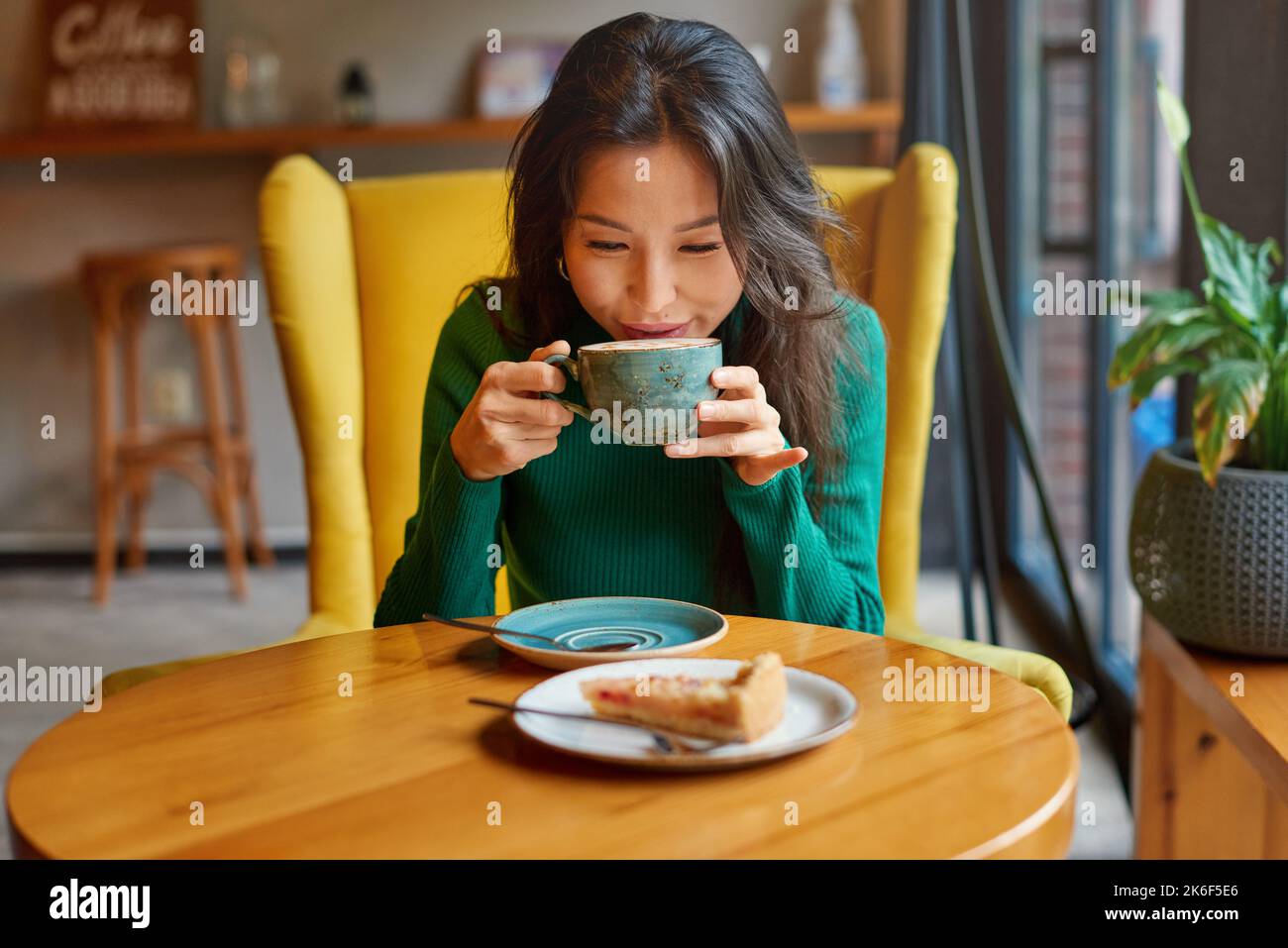 Ritratto di giovane asiatico affascinante, attraente brunetta kazako donna bere caffè caldo con torta di mele a tavola, seduta in accogliente sedia gialla in caffè. Foto Stock