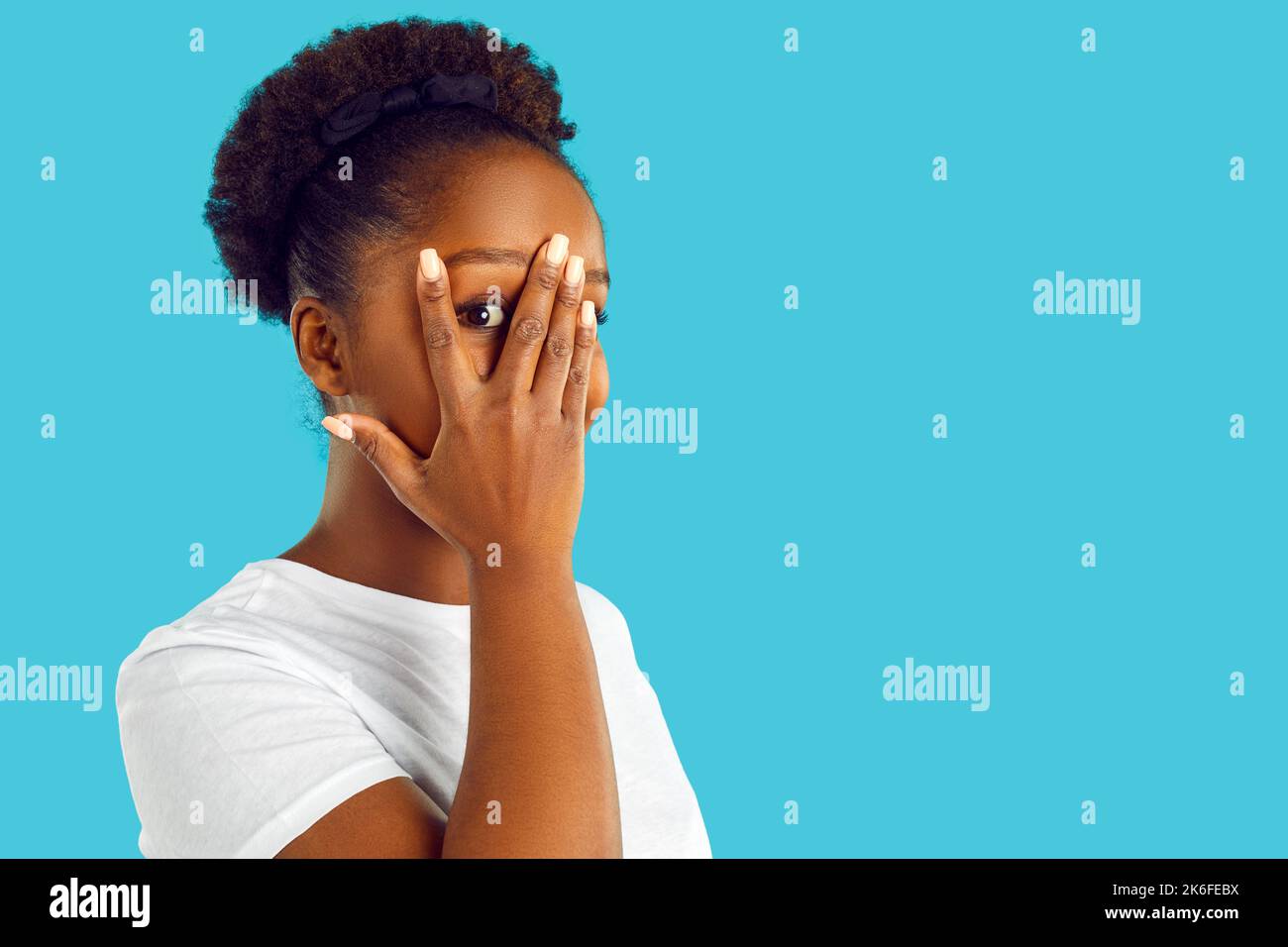 Timida giovane donna che copre il suo volto e guardando attraverso le dita su sfondo blu copia spazio Foto Stock
