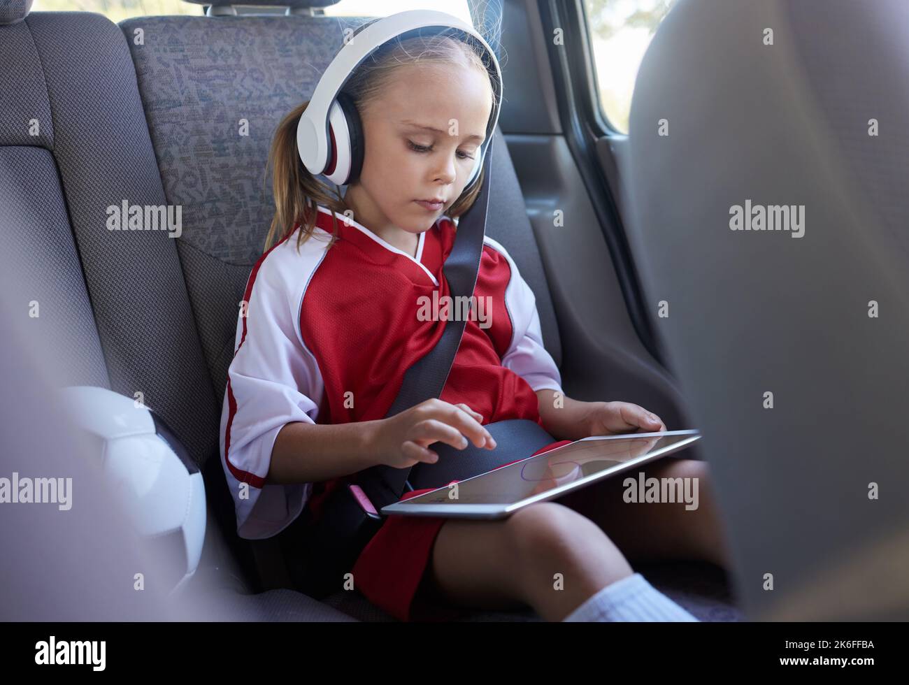 Bambino con tablet, cuffie e cintura di sicurezza in auto che tornerà a casa dopo una partita di calcio, una partita o una pratica. Ragazza giovane in sport gioco di attrezzature Foto Stock