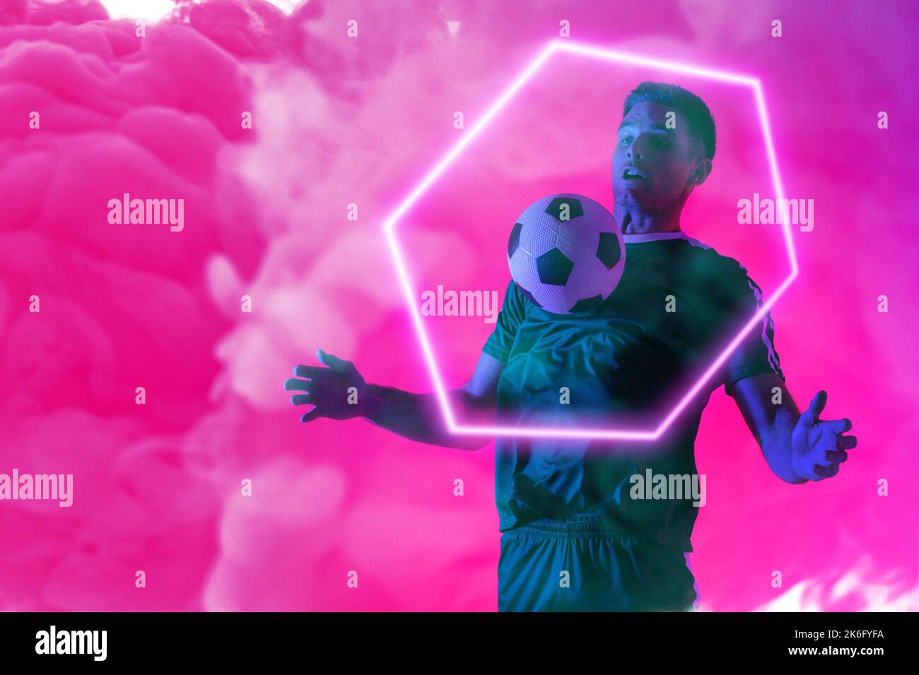 Giocatore maschio caucasico che gioca con la palla da calcio sopra l'esagono illuminato contro il fumo al neon rosa Foto Stock