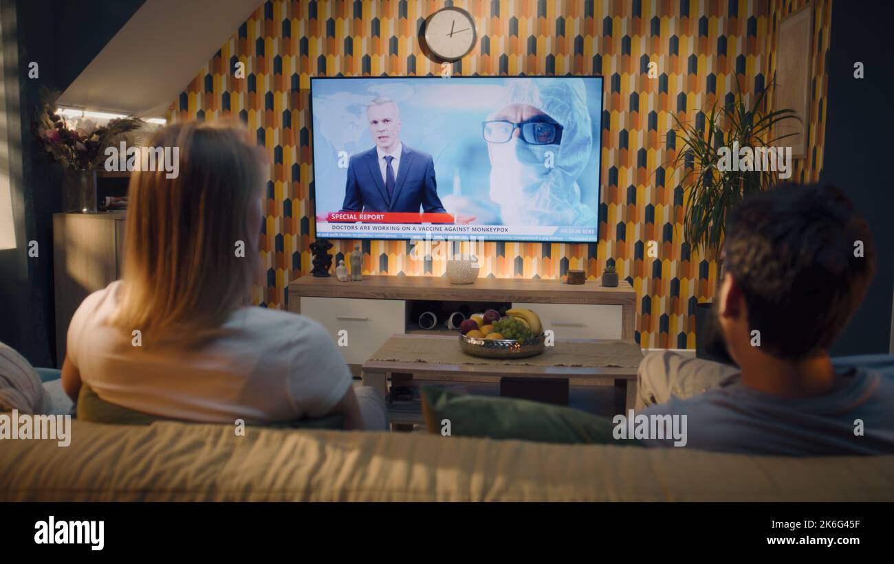 Coppia seduta sul divano insieme nel soggiorno con design moderno, guardando la medicina ultime notizie in TV sul vaiolo moneypox e lo sviluppo di vaccini. I coniugi trascorrono il fine settimana e si raffreddano a casa. Foto Stock