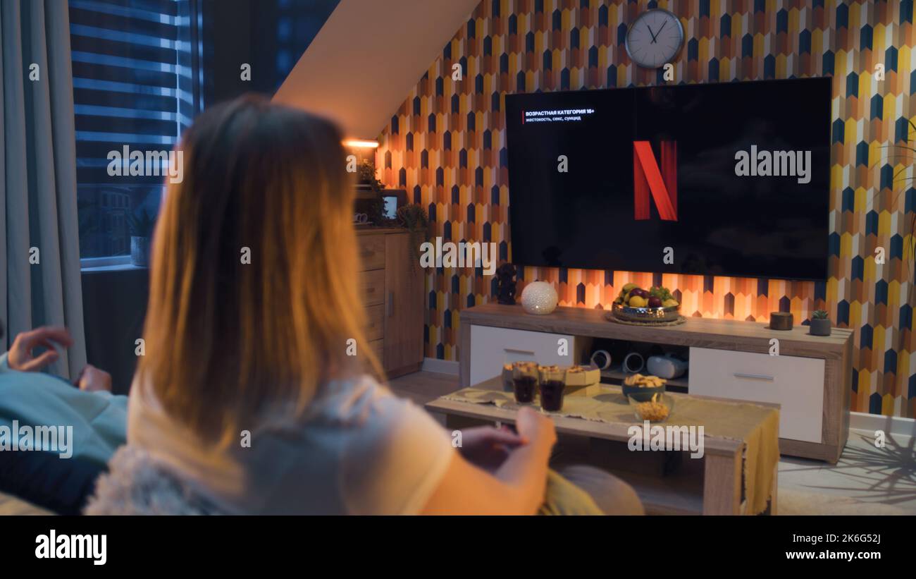 ZAPORIZHYA, UCRAINA - 13 ottobre 2022: Coppia seduta sul divano insieme, guardando film o serie TV su Netflix servizio di streaming con snack, bibite e pizza. L'uomo e la donna che si raffreddano riposano a casa. Foto Stock