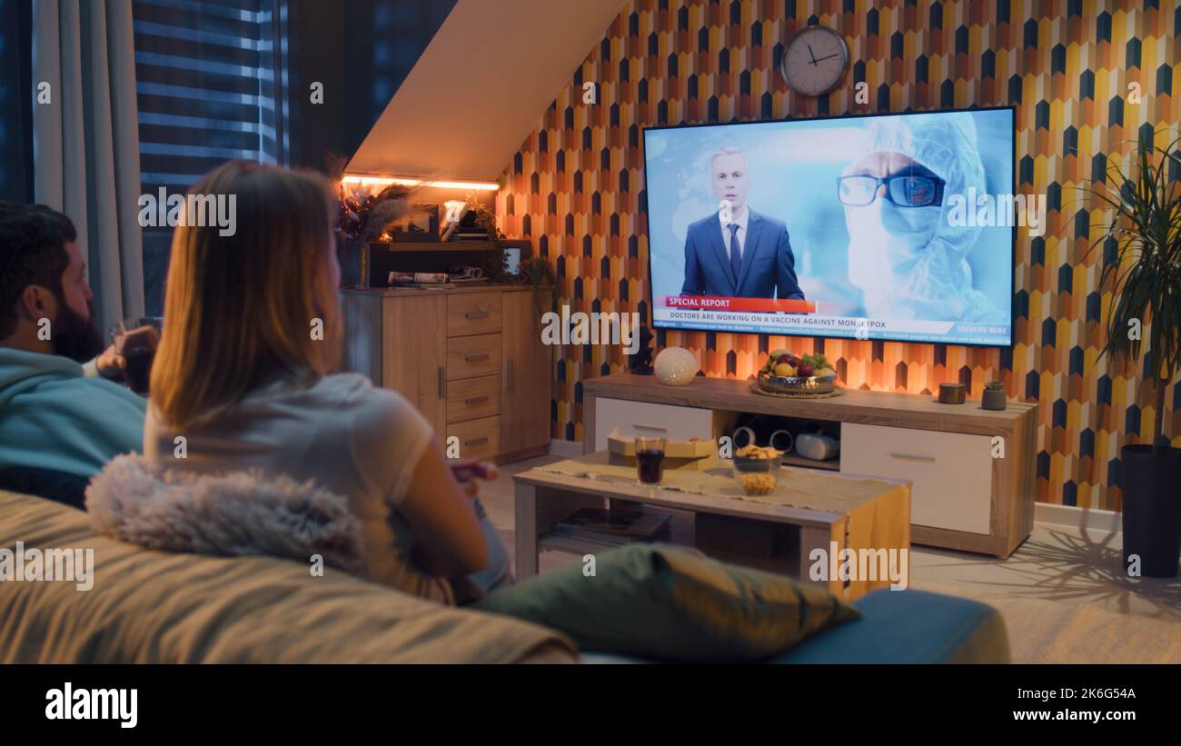 Coppia seduta sul divano insieme nel soggiorno con design moderno, guardando la medicina ultime notizie in TV sul vaiolo moneypox e lo sviluppo di vaccini. I coniugi trascorrono il fine settimana e si raffreddano a casa. Foto Stock