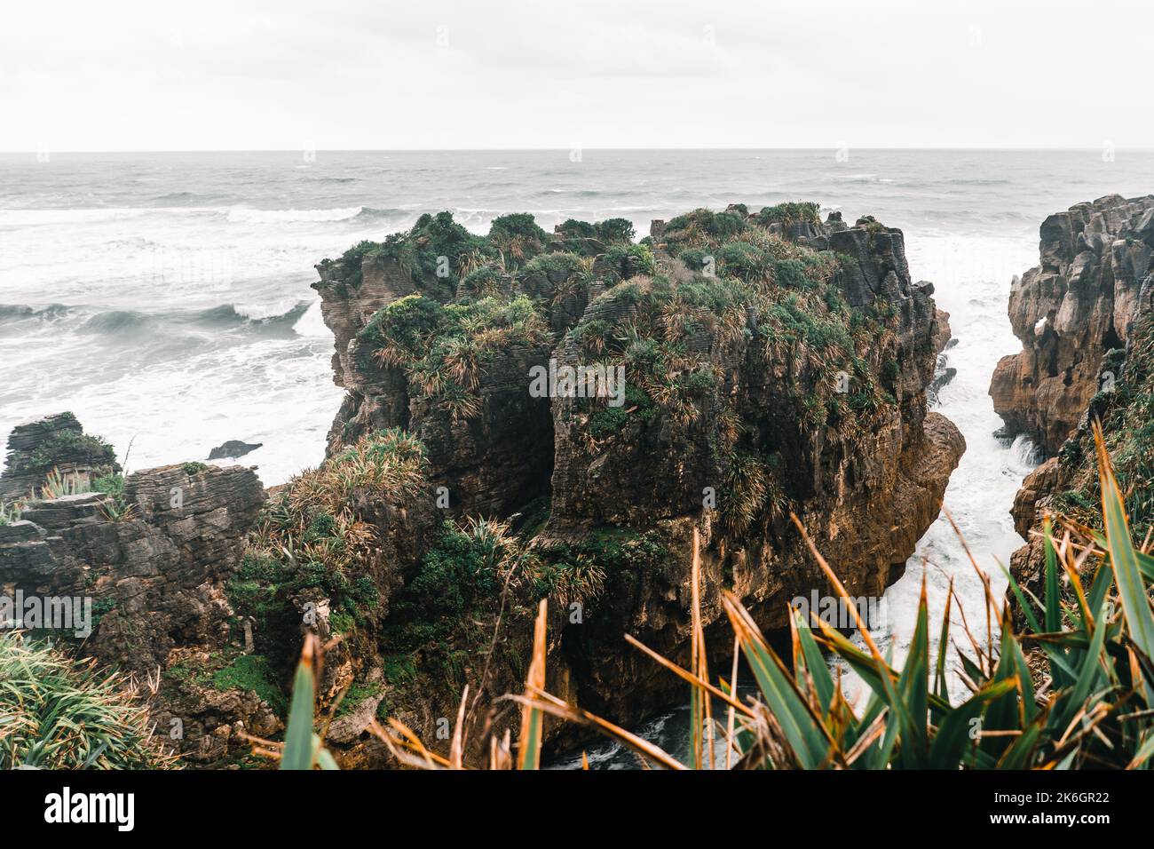 rocce strane con vegetazione e piante in cima alla spiaggia vicino ad un mare con molte onde al giorno con il maltempo e molte nuvole all'orizzonte Foto Stock
