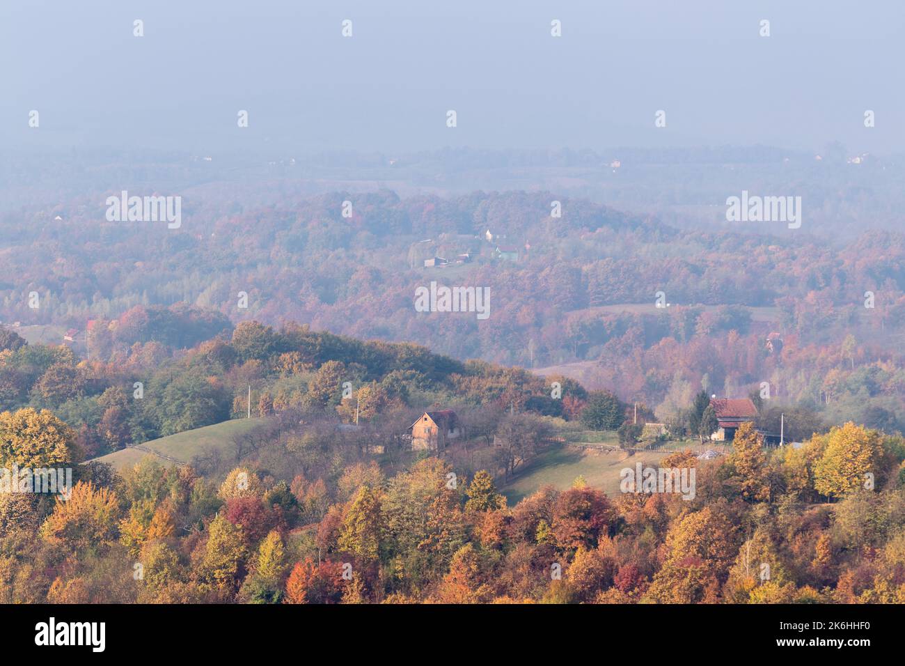 Case di villaggio sulla collina circondata da una lussureggiante foresta di colori autunnali, colline lontane svaniscono nella nebbia mattutina Foto Stock