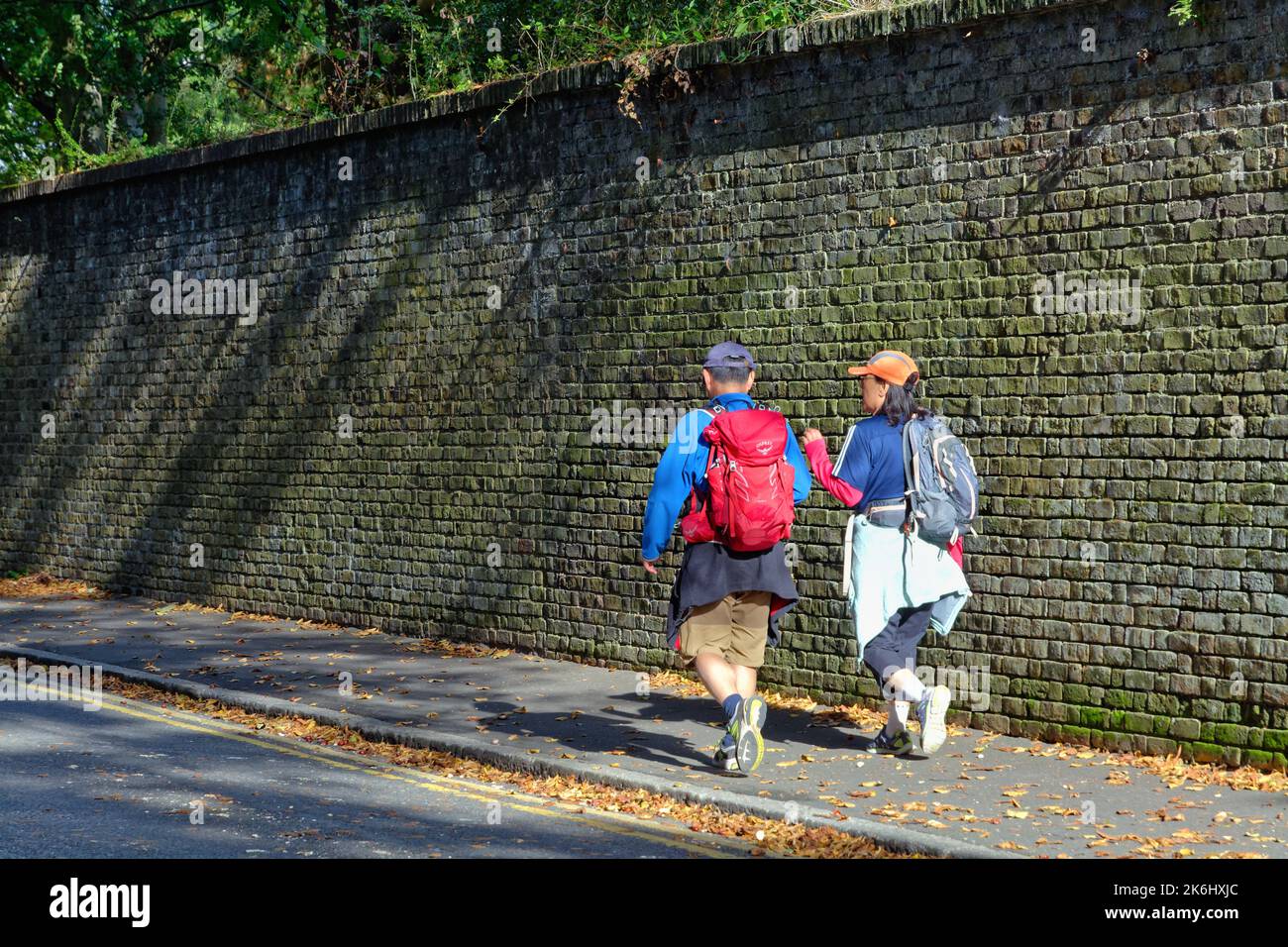 Coppia orientale di mezza età che indossa un abito a piedi su un marciapiede con un grande muro di mattoni sullo sfondo, Shepperton Surrey Inghilterra UK Foto Stock