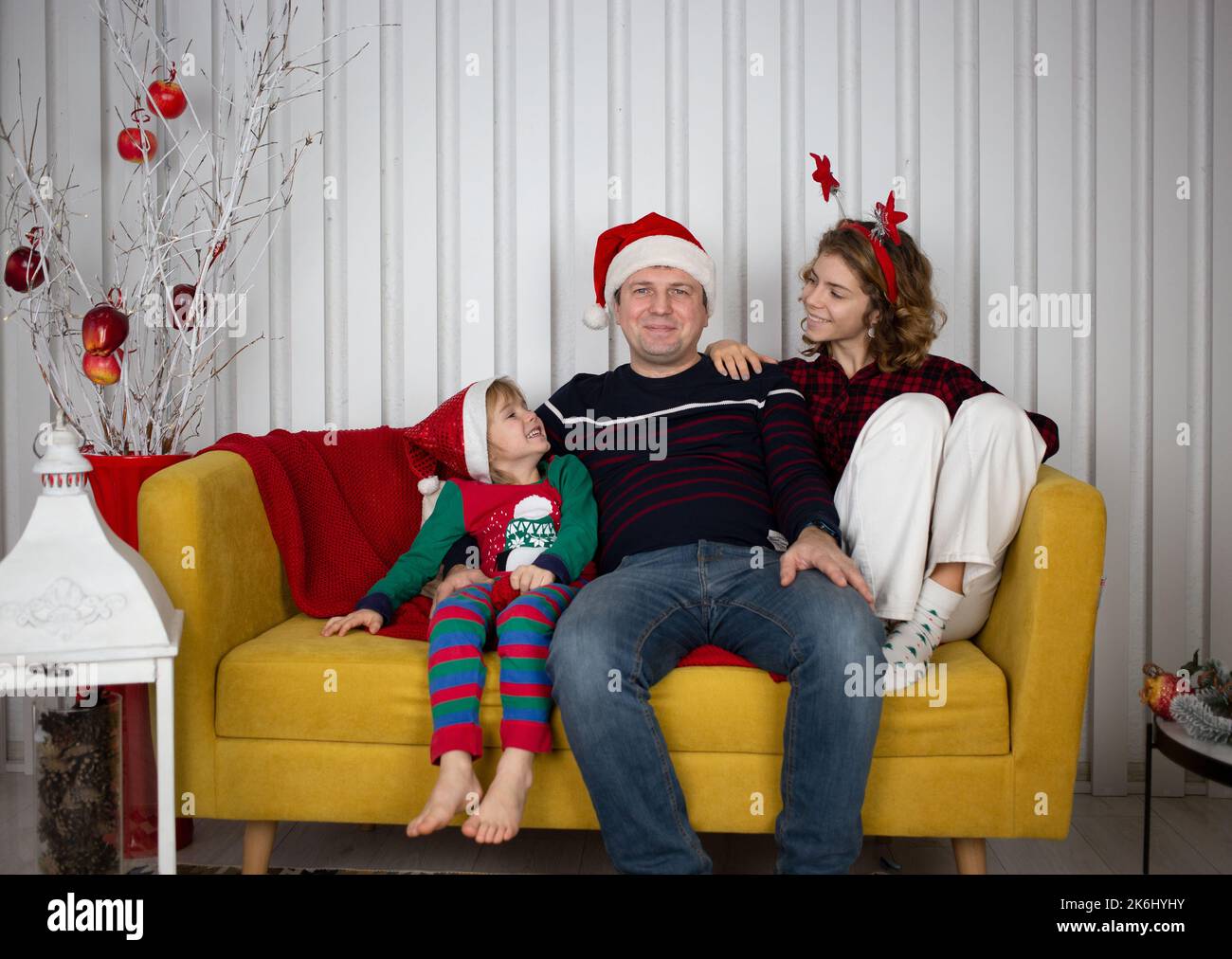 Felice anno nuovo e Buon Natale. papà gioioso e i suoi figli sono seduti in camera su un luminoso divano giallo in cappelli di santa. emozioni positive e amichevoli Foto Stock