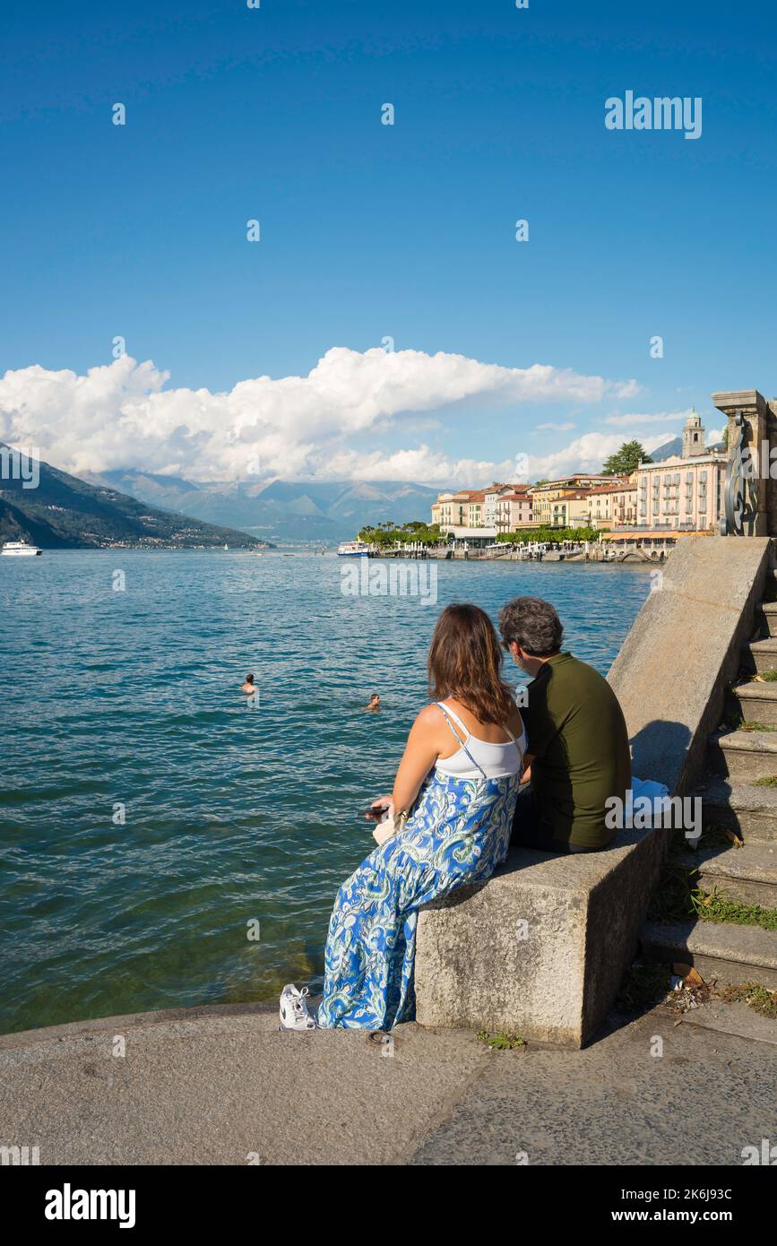 Laghi italiani, vista posteriore di una coppia di mezza età in estate seduti insieme sui gradini del lago a Bellagio e guardando i nuotatori sul lago di Como Foto Stock