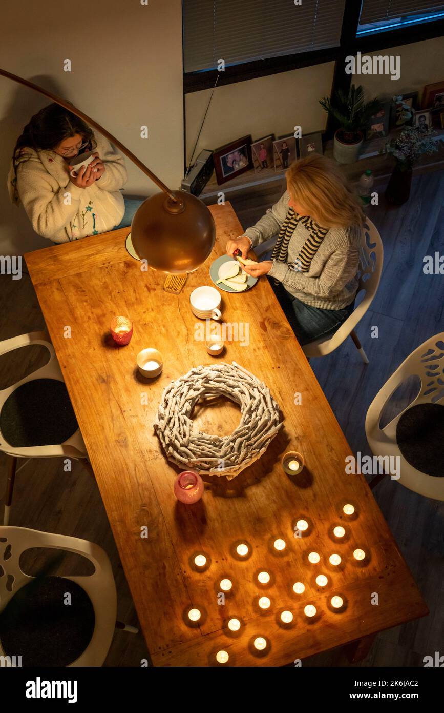 Immagine simbolica risparmio energetico, inverno freddo, crisi energetica, piatto freddo, madre e figlia vestite calorosamente a cena, una sola lampada accesa, a lume di candela, caldo Foto Stock