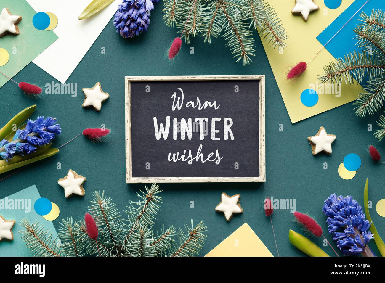 Testo di saluto caldo inverno auguri su lavagna, gesso tavola. Sfondo di carta geometrico verde con fiori di giacinto blu su carta colorata stratificata. Foto Stock