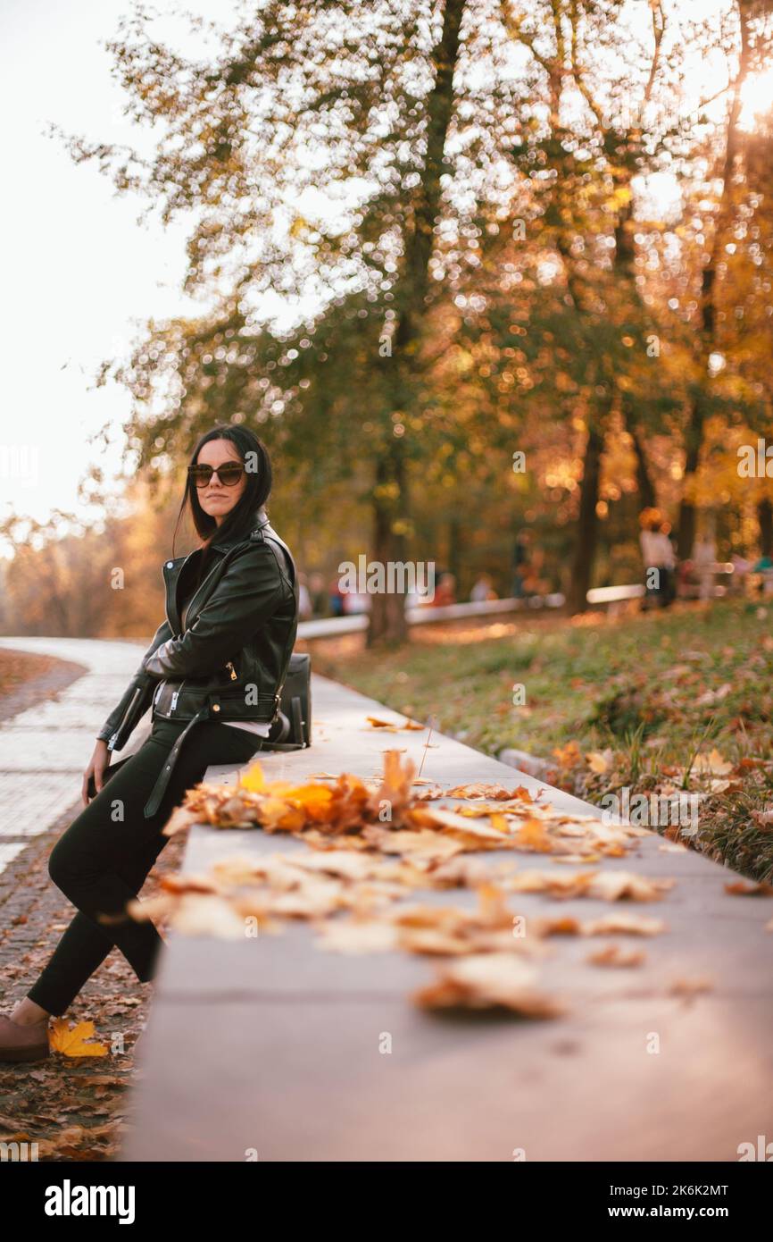 Ritratto di giovane donna elegante che indossa occhiali da sole e giacca in pelle in piedi nel parco durante il sole in autunno Foto Stock