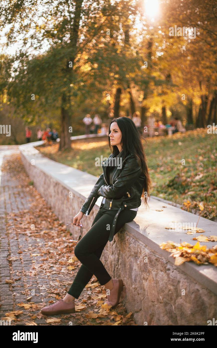 Ritratto di donna triste e premurosa che indossa una giacca in pelle in piedi nel parco durante il sole in autunno Foto Stock