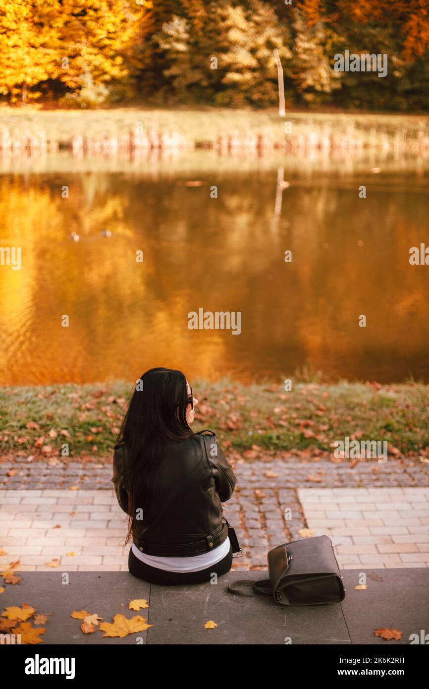 Vista posteriore della giovane donna alla moda pensierosa che guarda lontano mentre si siede vicino al lago nel parco durante il tempo soleggiato in autunno Foto Stock