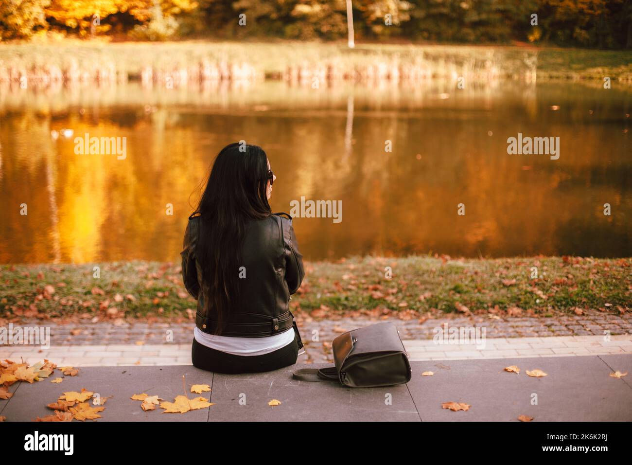 Vista posteriore della giovane donna alla moda pensierosa che guarda lontano mentre si siede vicino al lago nel parco durante il tempo soleggiato in autunno Foto Stock