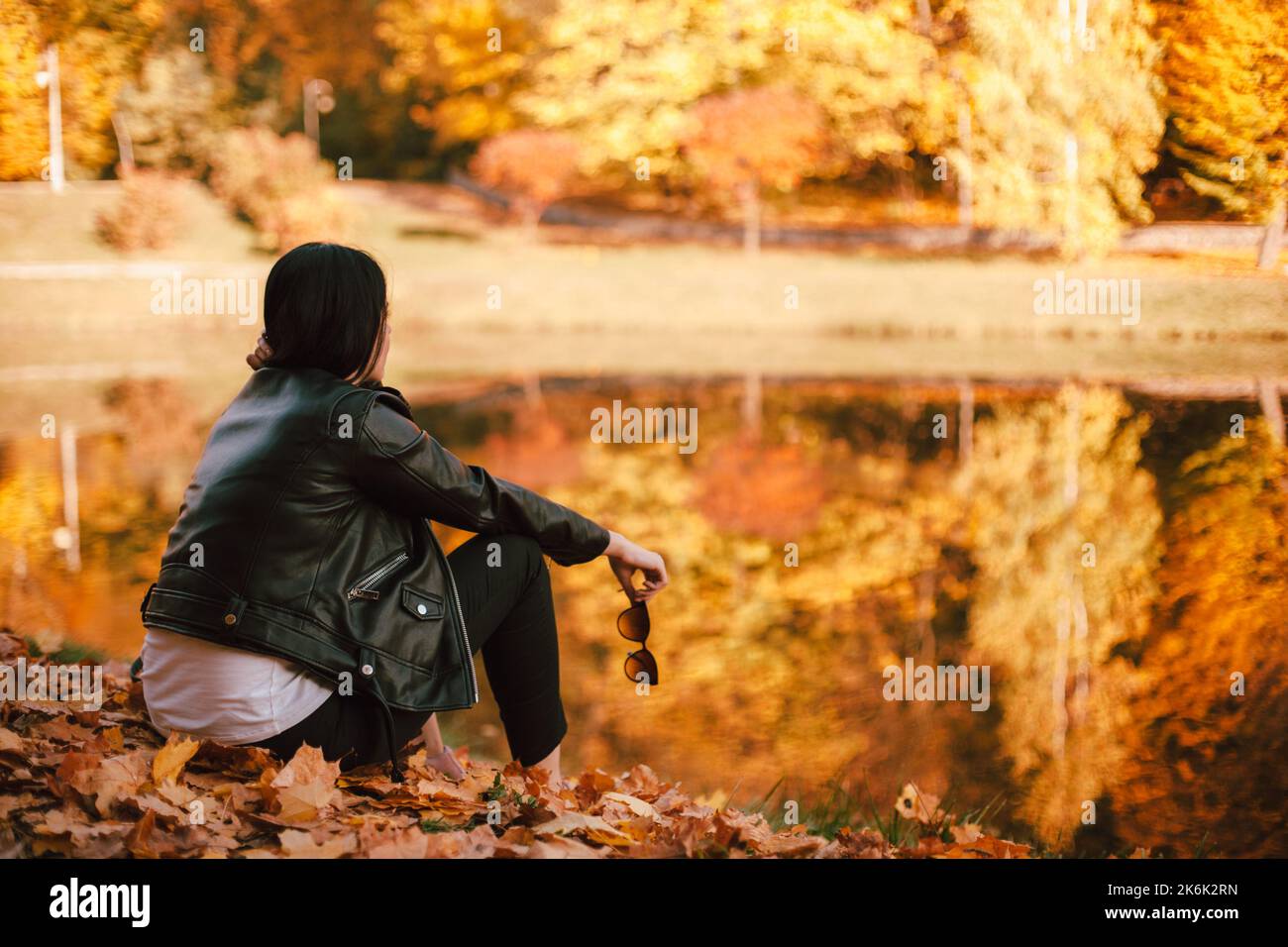 Giovane donna attenta ed elegante che guarda lontano mentre si siede vicino al lago nel parco durante il tempo soleggiato in autunno. Alberi di arancio riflessi nell'acqua Foto Stock