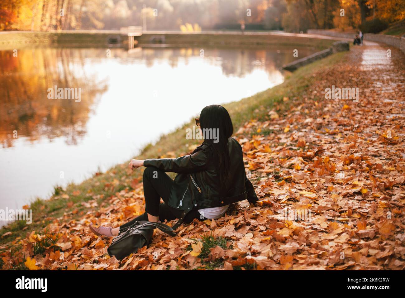 Giovane donna elegante e tranquilla che guarda la vista panoramica mentre si siede sulle foglie cadute dal lago nel parco durante il sole in autunno Foto Stock