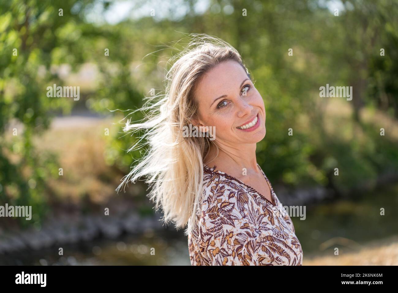 Donna bionda di mezza età che guarda giocosamente sopra la spalla con un sorriso di fascio all'aperto nella natura nel verde Foto Stock