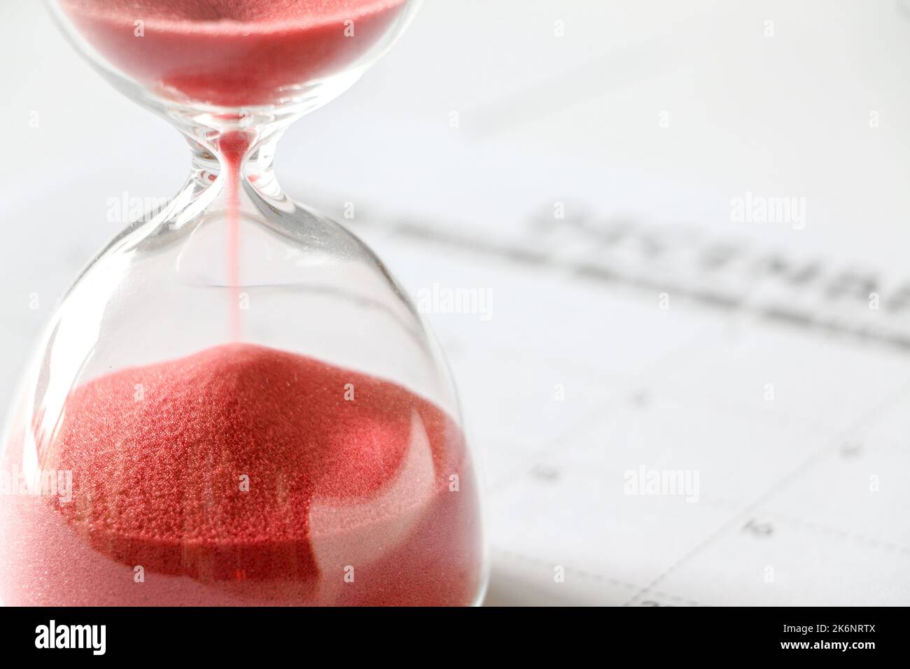 Primo piano di sabbia rossa che scivola via attraverso la clessidra, tempo che sta per scadere, calendario di dicembre fuori fuoco in background, fine dell'anno, non più tempo Foto Stock
