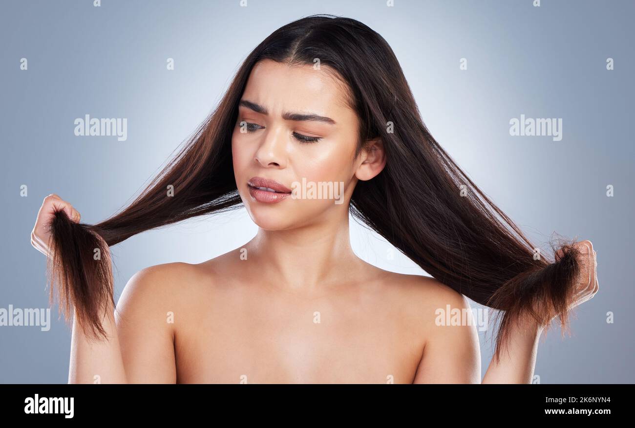 Giovane donna di razza mista che tiene i suoi lunghi capelli castani e guarda le doppie punte danneggiate. Donna che guarda turbato circa i problemi di cura dei capelli Foto Stock