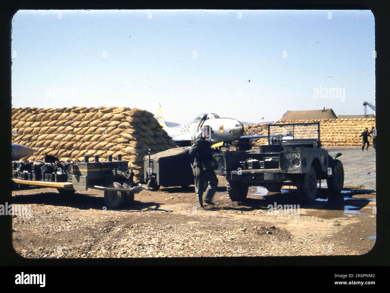 Vista frontale a metà destra di una Lockheed F-80 Shooting Star parcheggiata in una rivaccata, da qualche parte in Corea. Il numero di ronzio, FT-858, è visibile sul lato. Una jeep è parcheggiata in primo piano e un uomo si trova accanto ad essa e porta qualche attrezzatura. Altri veicoli di supporto sono parcheggiati nelle vicinanze. Uomini, attrezzature ed edifici appaiono sullo sfondo. Foto Stock