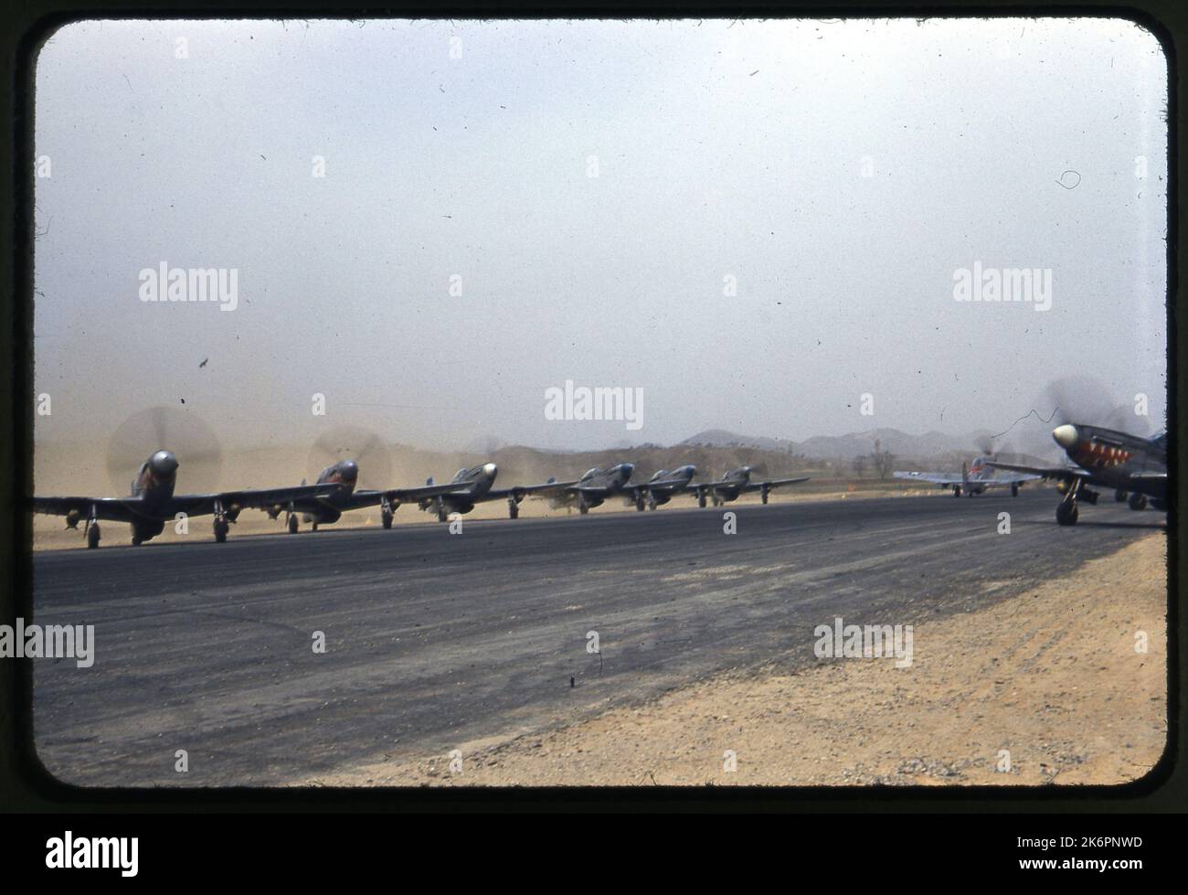Vista posteriore destra di tre quarti di un F-51D Mustang tassante nordamericano, da qualche parte in Corea. Accanto alla pista sul lato sinistro dell'immagine è parcheggiata una linea di volo di F-51D Mustangs. Un altro F-51D Mustang è parcheggiato sul lato destro della pista. Tre degli aerei portano le marcature del 12th Fighter Bomber Squadron (FBS). Ogni aereo è armato di due bombe. Foto Stock