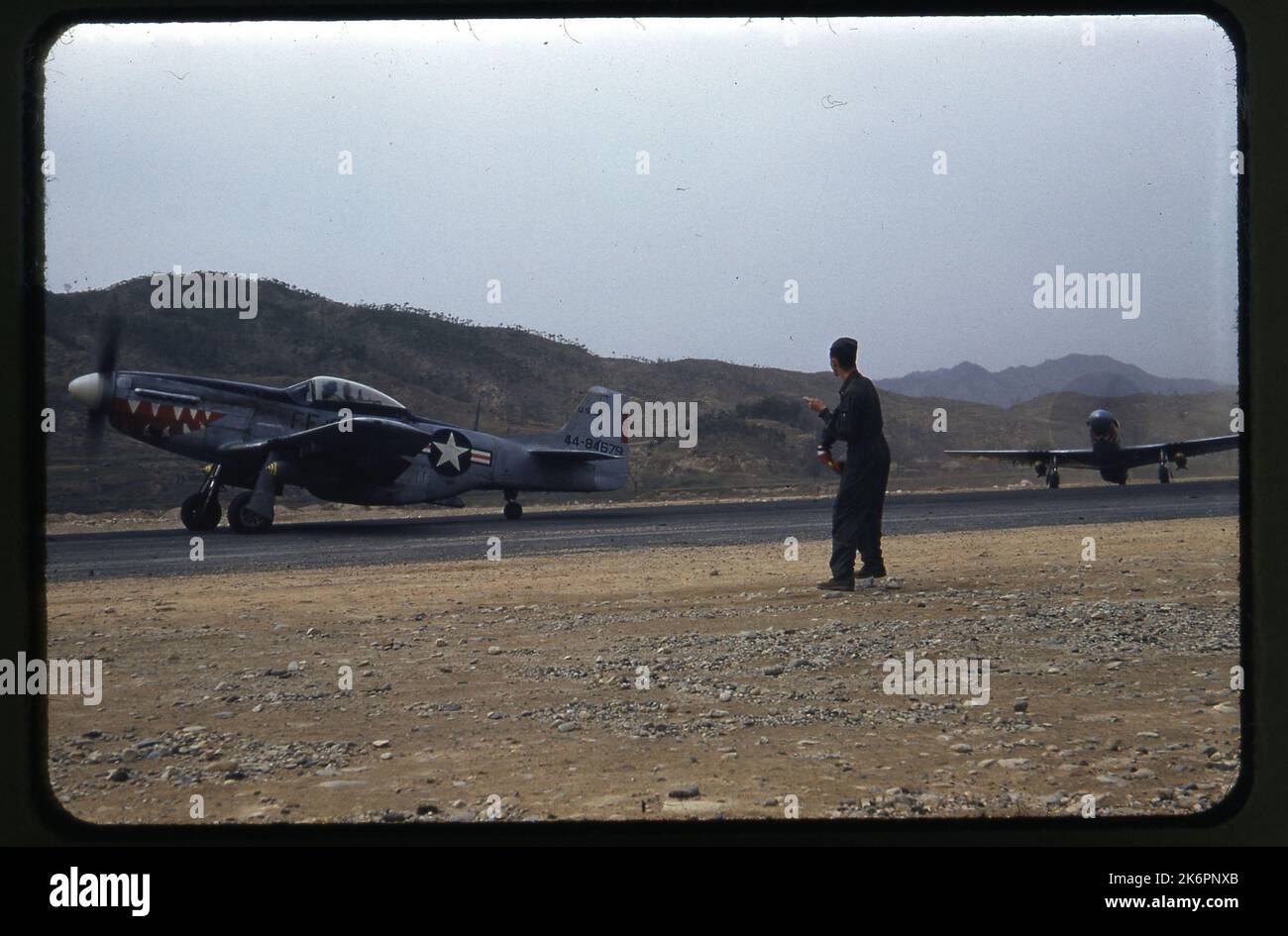 Un quarto della vista anteriore sinistra di un F-51D Mustang nordamericano (s/n 44-84679; c/n 44535) che tassa, in qualche luogo in Corea. L'aereo è seguito lungo la pista da un altro F-51D Mustang. Entrambi portano le marcature del 12th Fighter Bomber Squadron (FBS), ed entrambi sono armati di due bombe. Un crewman si trova in primo piano nell'immagine. Foto Stock