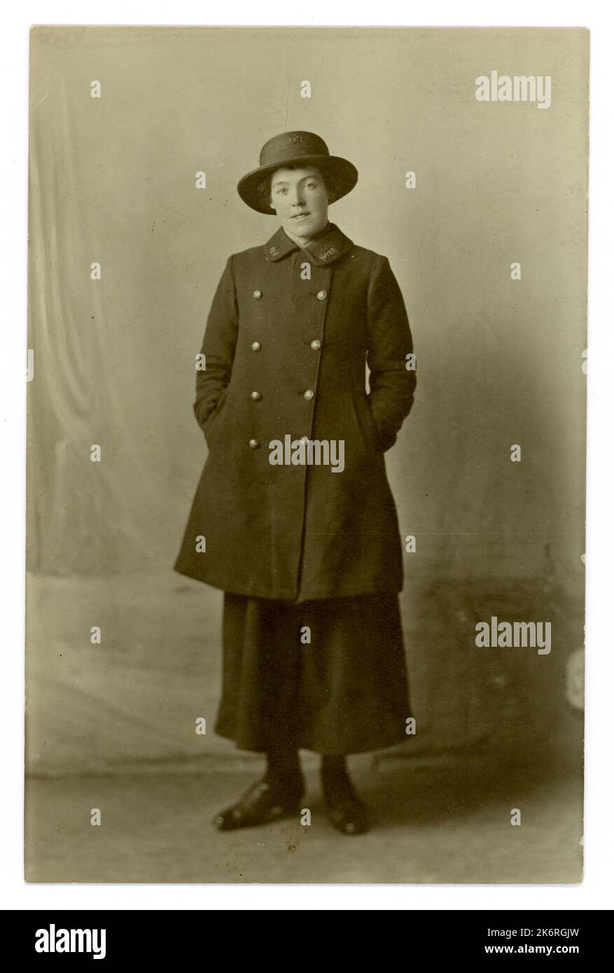WW1 era cartolina ritratto di lavoratrice postale femminile in uniforme di gonna di serge blu, cappotto e cappello di paglia blu. General Post Office (GPO) Poster / postwomen - aiutare a consegnare le lettere per aiutare lo sforzo di guerra sul fronte casa, circa 1916 studi Herman's, Wood Green, Londra, Regno Unito Foto Stock
