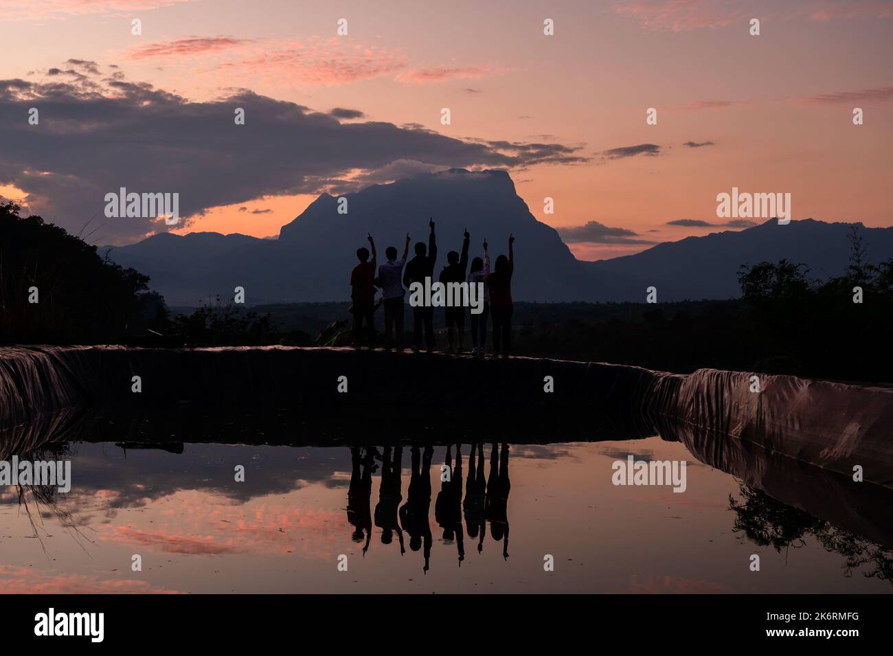 Gruppi di turisti in piedi con le braccia che puntano verso il cielo e riflettono la montagna nel lago al colorato cielo tramonto Foto Stock