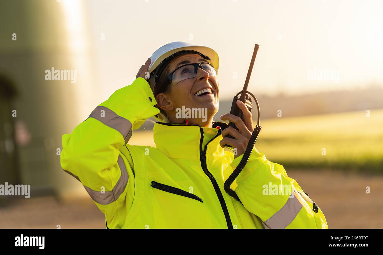 ingegnere donna con giacca di sicurezza in piedi davanti a una turbina eolica che parla in un walkie talkie al controllo del sistema di turbina eolica Foto Stock